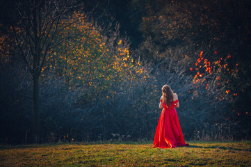 Mysterious sorceress in a beautiful red dress. Her hair and dress are fluttering in the wind. Background bright, autumn, fiery forest trees with warm tones.