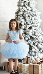 Little girl near fir-tree with Christmas gifts in white and gold holiday interior. Christmas celebration. Child Girl weared in blue dress near a New Year's gifts on the floor. New year eve