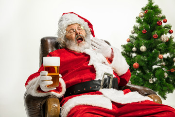 Santa Claus drinking beer near the Christmas tree, congratulating, looks drunk and happy. Caucasian male model in traditional costume. New Year 2020, gifts, holidays, winter mood. Copyspace for your