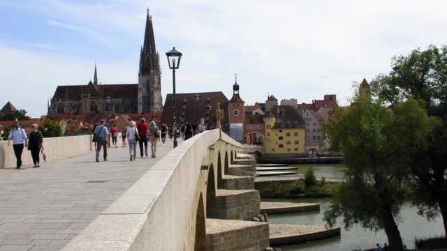 People walk along an old medieval stone bridge over the Danube River in the city of Regensburg Germany on a warm summer day. Historic buildings are visible in the background.