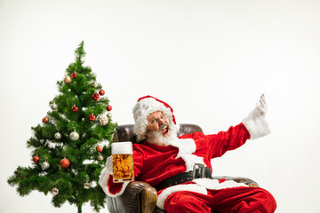 Santa Claus drinking beer near the Christmas tree, congratulating, looks drunk and happy. Caucasian male model in traditional costume. New Year 2020, gifts, holidays, winter mood. Copyspace for your
