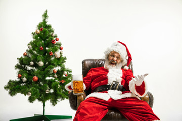 Santa Claus drinking beer near the Christmas tree, congratulating, looks drunk and happy. Caucasian male model in traditional costume. New Year 2020, gifts, holidays, winter mood. Copyspace for your