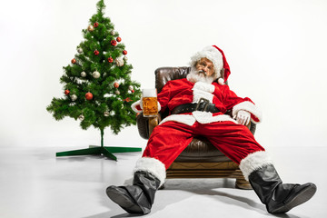 Santa Claus drinking beer near the Christmas tree, congratulating, looks drunk and happy. Caucasian male model in traditional costume. New Year 2020, gifts, holidays, winter mood. Copyspace for your