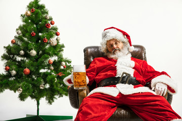 Fototapeta na wymiar Santa Claus drinking beer near the Christmas tree, congratulating, looks drunk and happy. Caucasian male model in traditional costume. New Year 2020, gifts, holidays, winter mood. Copyspace for your