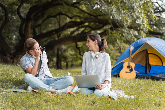 Young couples sit in the grassy field and take pictures and play computer, happily resting while camping in the midst of nature and beautiful trees.