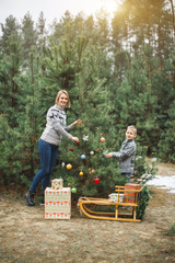 Mother and little son looking at camera and smiling, while decorate Christmas tree in winter forest, outdoors. New Year decorations, wooden sledge, present gifts