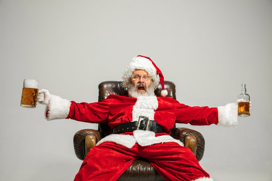 Santa Claus drinking beer sitting on armchair, congratulating, looks drunk and happy. Caucasian male model in traditional costume. New Year 2020, gifts, holidays, winter mood. Copyspace for your ad.