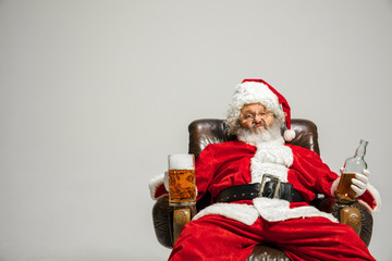 Santa Claus drinking beer sitting on armchair, congratulating, looks drunk and happy. Caucasian male model in traditional costume. New Year 2020, gifts, holidays, winter mood. Copyspace for your ad.