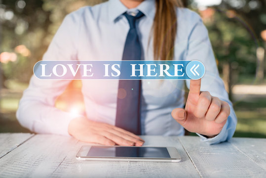 Handwriting text Love Is Here. Conceptual photo Roanalysistic feeling Lovely emotion Positive Expression Care Joy Female business person sitting by table and holding mobile phone