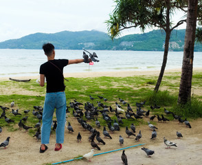 A young guy, in a black t-shirt and blue jeans, in flip-flops of different colors, feeds pigeons...