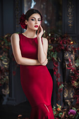 Fototapeta na wymiar Attractive gorgeous brunette woman in red evening dress in dark luxury interior in vintage barocco style decorated flowers