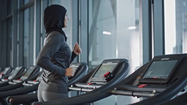 Athletic Muslim Sports Woman Wearing Hijab and Sportswear Running on Treadmill. Energetic Fit Female Athlete Training in Gym Alone. Urban Business District Window View. Side View Portrait Slow Motion
