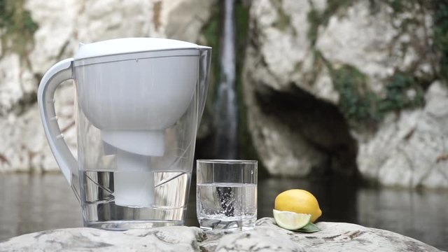 Filter jug and transparent glass of a clear water with lemon open air on the background of a mountain stream and waterfall