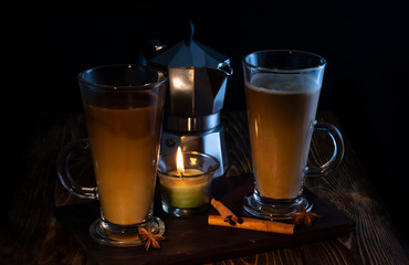 Two glasses of coffee with a coffee maker in the background on a wooden table with chocolate, anise, karatz lit by candle flame.
