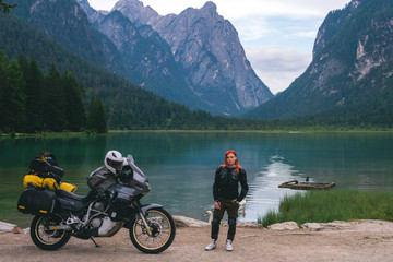 Girl motobiker in protective turtle jacket stands on the beach with tourer motorcycle. Extreme travel vacation, motorcyclist adventure lifestyle. Toblacher See, (Italian: Lago di Dobbiaco) Italy.