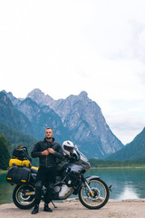 Handsome man motorcyclist with touring motorcycle on beach. Alpine mountains and lake on background. Biker lifestyle, traveler. Toblacher See, (Lago di Dobbiaco) Italy. leather jacket, vertical photo