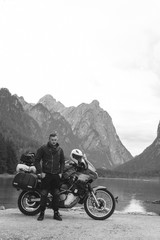 Handsome man motorcyclist with touring motorcycle on beach. Alpine mountains and lake on background. Biker lifestyle, traveler. Toblacher See, (Lago di Dobbiaco) Italy. black and white vertical