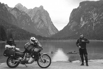 Handsome man motorcyclist with touring motorcycle on beach. Alpine mountains and lake on background. Biker lifestyle, traveler. Toblacher See, (Lago di Dobbiaco) Italy. copy space, black and white