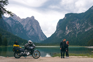 Motorcyclists couple stand on beach with touring notorcycle. Lake and Alpine mountains on background. Destination concept. Happy together. Copy space. Toblacher See, (Lago di Dobbiaco) Italy