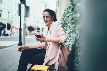 Happy blogger making payment online on smartphone device via 4G internet resting on bench outdoors, successful young woman in stylish jacket and glasses messaging in friendly chat on mobile phone