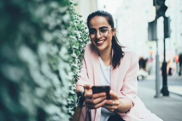 Half length portrait of pretty cheerful woman in glasses laughing during sending joke messages to friends via smartphone application, happy traveler looking at camera during having fun in network