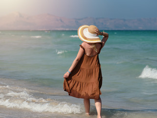 Fototapeta na wymiar Young blond European woman in colorful sun hat standing against wonderful blue sea. She enjoying her rest at seacoast and looking towards sea.