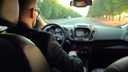 Formally dressed bearded man in glasses driving a car in sunny weather. View from the back seat of a car