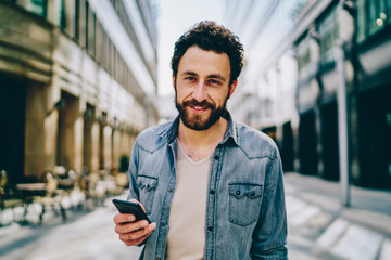 Portrait of handsome young man with beard smiling at camera while dialing number on phone standing...