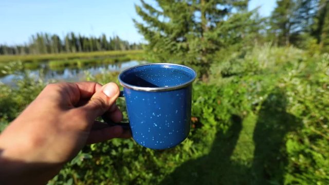 A hand holds a blue enamel coffee cup in the sun. The camera pans with the cup revealing the natural scenery in slow motion with selective focus