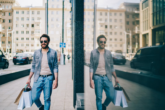 Handsome caucasian male buyer carrying bags with purchases satisfied with his job, happy hipster guy in trendy clothes and sunglasses walking near store in downtown after shopping enjoy free time.
