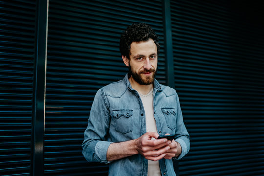 Half length portrait of handsome caucasian man holding mobile phone standing on street in casual wear, hipster guy typing message on modern smartphone chatting online via 4G internet connection.