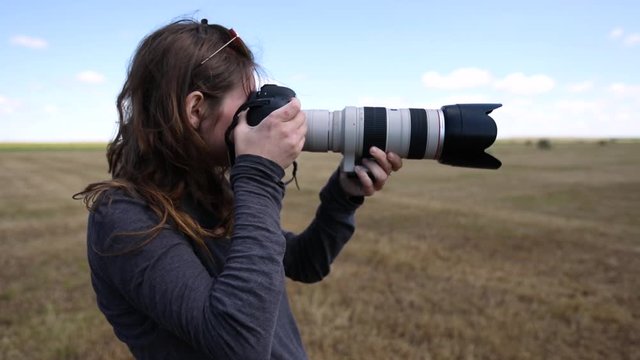 A female photographer uses a professional camera and telephoto lens to capture the landscape. She stands in a field and is seen in slow motion