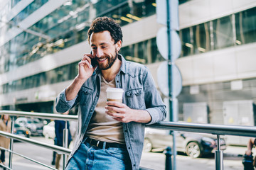 Cheerful bearded hipster guy dressed in stylish casual wear laughing during conversation on smartphone device.Positive young man with coffee to go in hand resting and talking on mobile on balcony