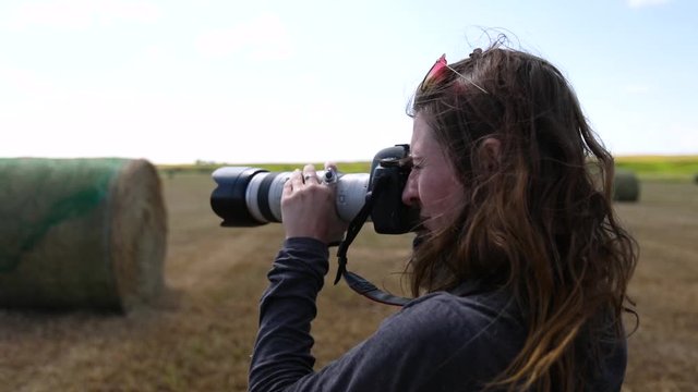 A female photographer stands in a field with her camera to her eye. She uses a telephoto lens to photograph a haystack and is seen in slow motion
