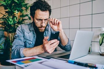 Serious guy in eyewear taking off eyewear while read carefully message on mobile phone about banking payment requires, confused freelancer businessman checking news from exchange on smartphone.