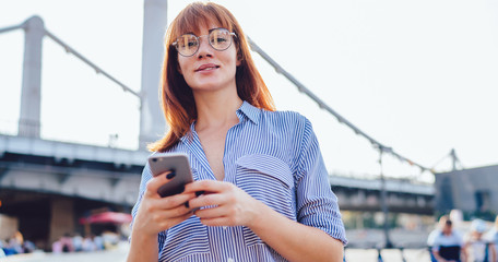 Half length portrait of attractive Caucasian female traveller with cellphone gadget in hands looking at camera while browsing web page with location information, concept of millennial time and tourism