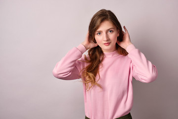 A girl with clean skin in a pink sweatshirt stands on a gray background and looking in the frame, collects hair. 