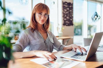 Portrait of attractive female manager of cafeteria working with online documents for organize plan, beautiful woman in eyeglasses looking at camera sitting at desk with modern laptop device - obrazy, fototapety, plakaty