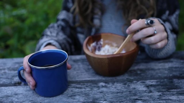 Woman eating breakfast outdoors in early morning. She warms her hand on her coffee cup while eating her muesli from a wooden bowl. 