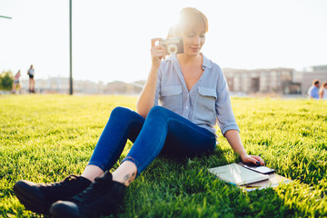Attractive female amateur checking notes about photography skills in textbook for education, beautiful Caucasian woman with retro camera in hands reading information from notepad sitting on park grass