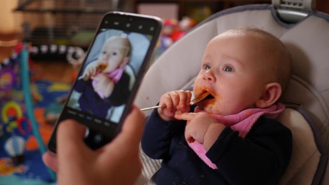 Mother takes phone picture of her baby child daughter smeared smudged face play with spoon after first eating nutrition vegetable puree instead of breath milk