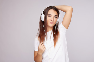 Headshot of young pleasant woman listening, enjoying music via online player, wearing white earphones, t-shirt, posing indoors against grey studio wall with copy space for text or advertising content