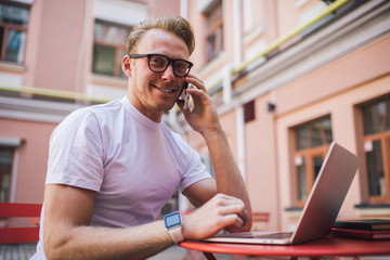 Portrait of happy male blogger in classic glasses using cellphone connection for phoning to best friend and talking about success with startup, skilled man sitting outdoors and smiling at camera