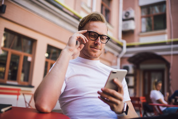 Positive male blogger reading received comments from followers near web publication about technology influence, successful hipster guy in spectacles updating profile in social networks via app