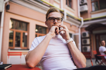 Pensive hipster guy in spectacles making international cellphone conversation sitting at urban setting and using roaming connection, contemplative man talking with customer service for consultancy