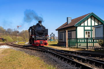 Dampflok, Schmalspurbahn bei einfahrt in Bahnhof mit Stellwerkhäuschen und Weichen vor blauem Himmel mit dampf