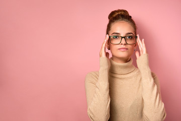 A girl with clean skin and a high bun stands on pink background and looks into the frame, adjusting her glasses with her fingers.