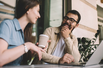 Selective focus on bearded asian mal listening to caucasian female friends talking while sitting in coffee shop next to laptop computer, positive young colleagues having conversation on work break.