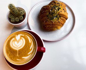 On a white table is a red cup of coffee, a bun with cheese and black sesame seeds, a cactus.