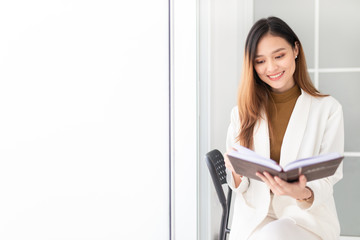 attractive asian female office reading a book in office, she sitting on seat and holding a book with hand, she feeling happy and smile, knowledge management